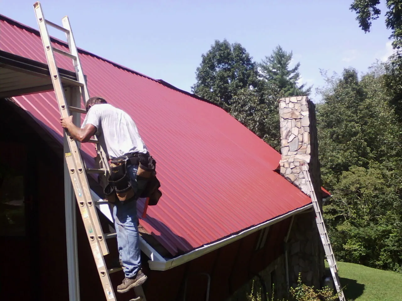 Roofer on a ladder beside a red metal roof and stone chimney — Soffit & Fascia Repair by Roofing Brothers in Humeston